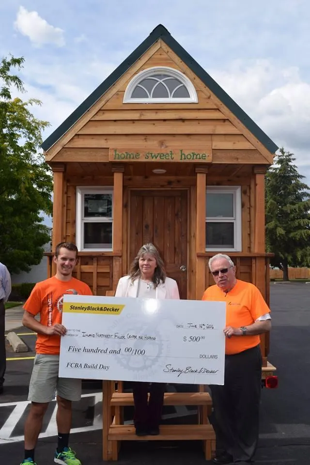 Two volunteers holding a giant donation check in front of tiny home