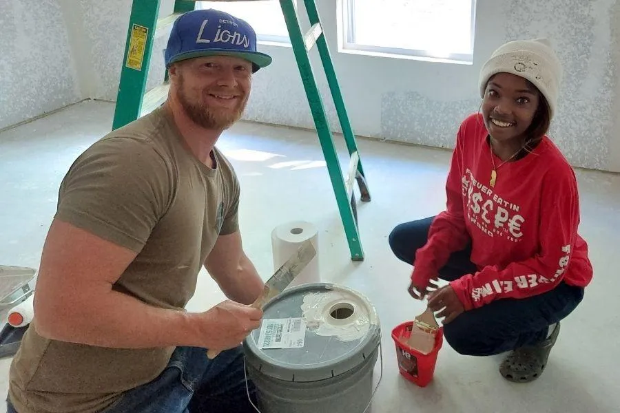 Two volunteers sitting on living room floors with paint buckets and rollers