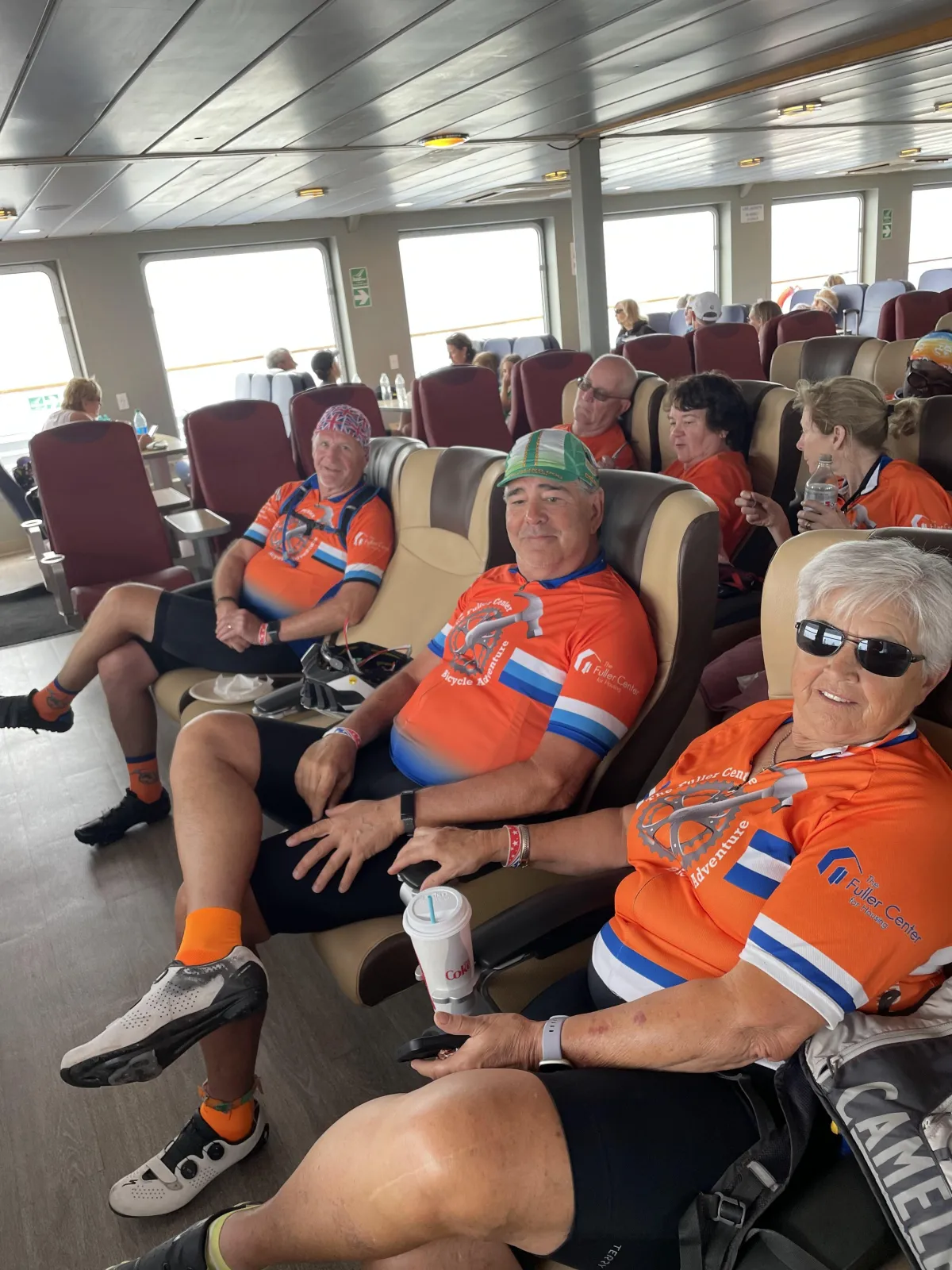 Cyclists sitting indoors in orange shirts, smiling and relaxing on he Lewes-Cape May Ferry.