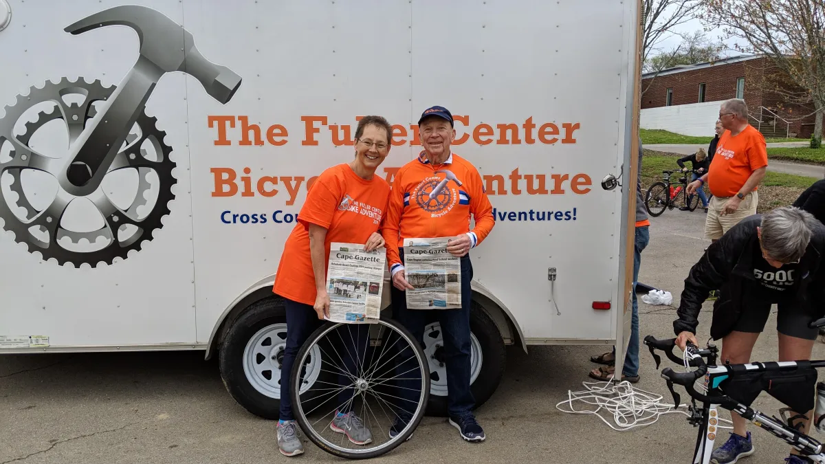 Michele and Jay smiling, holding Cape Gazenewspaper in front of the Fuller Center trailerte local 