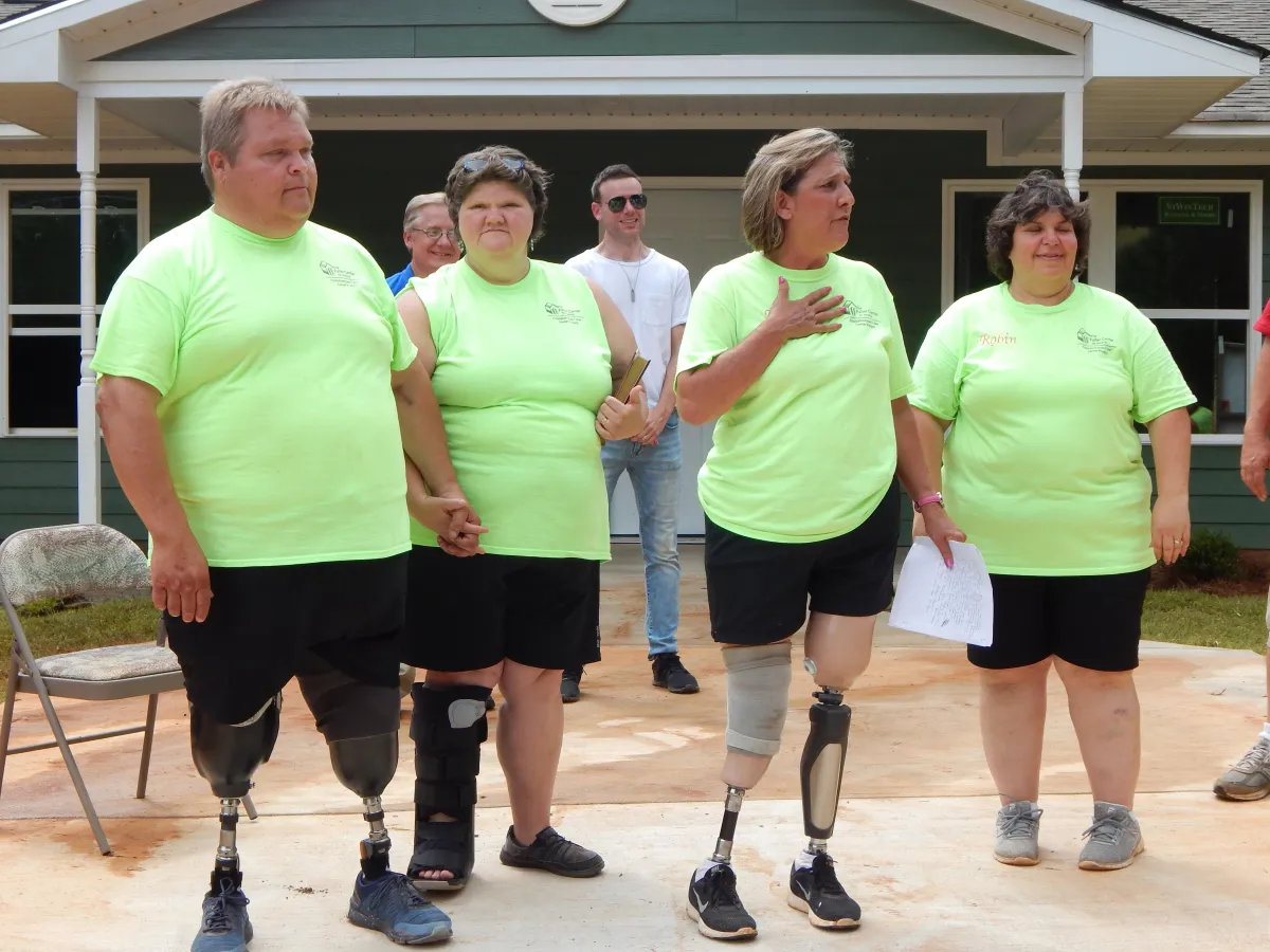 A group of Fuller Center volunteers and homeowners, including individuals with prosthetics, standing hand-in-hand during a heartfelt dedication in front of a newly repaired home.