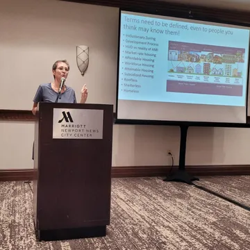 Dr. Tiny speaking at a podium in front of a presentation screen during a housing workshop.