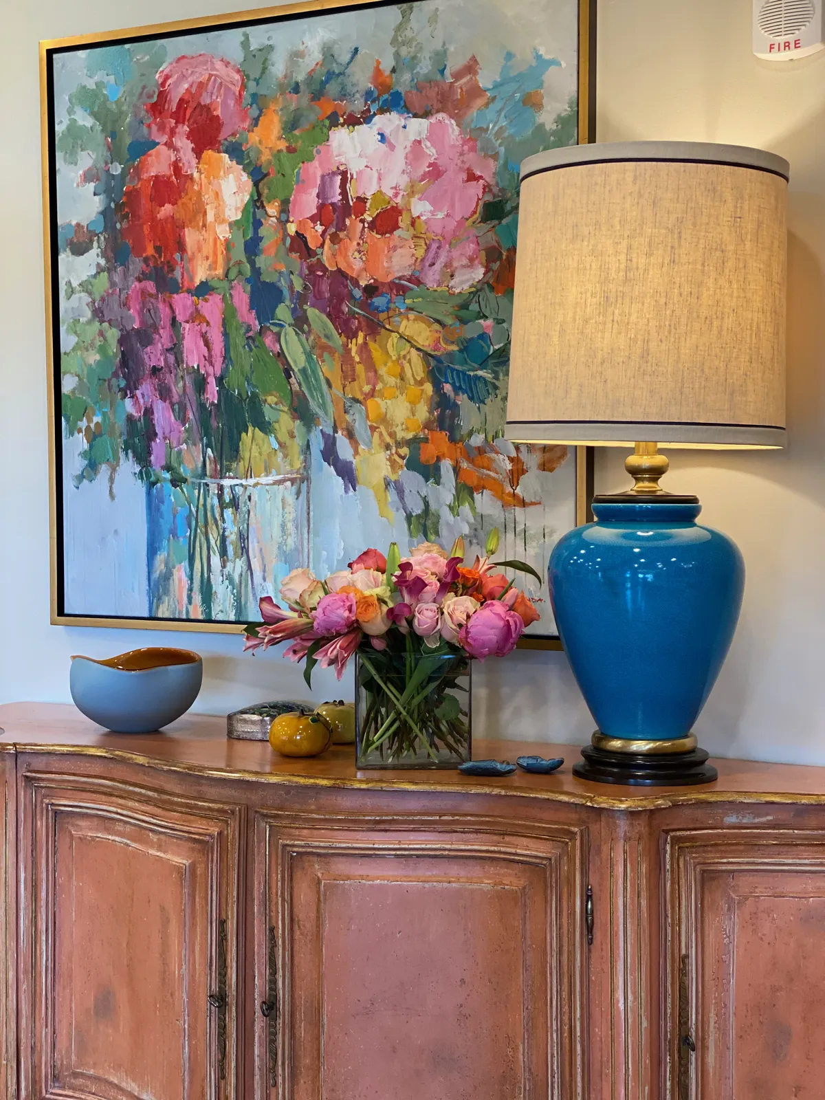 Floral painting above a pink sideboard with a blue ceramic lamp
