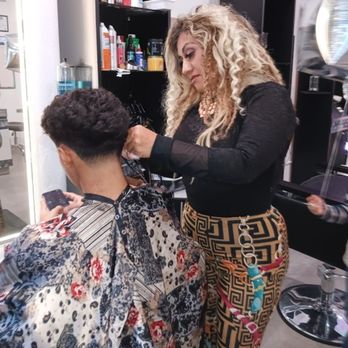 A smiling bilingual mother and daughter, both with stylish hair and manicured nails, stand together in a bright, modern salon interior with soft pink and gold accents. The background shows a glimpse of Fremont Street through the window, capturing the vibrant Las Vegas atmosphere. The image is warm, inviting, and professional, emphasizing their approachable and personal touch.