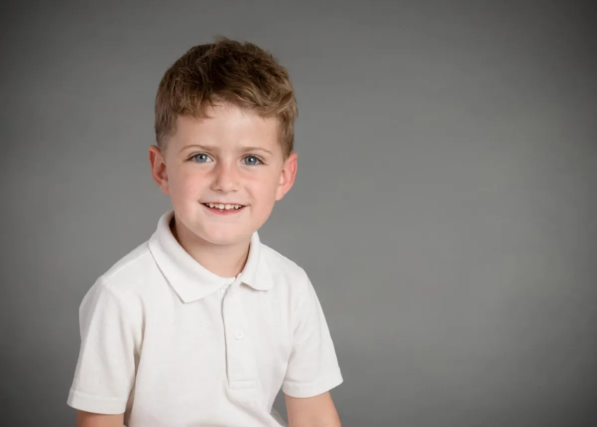 Portrait of a happy young boy with blonde hair, blue eyes, and playful smile, seated against a neutral studio background.
