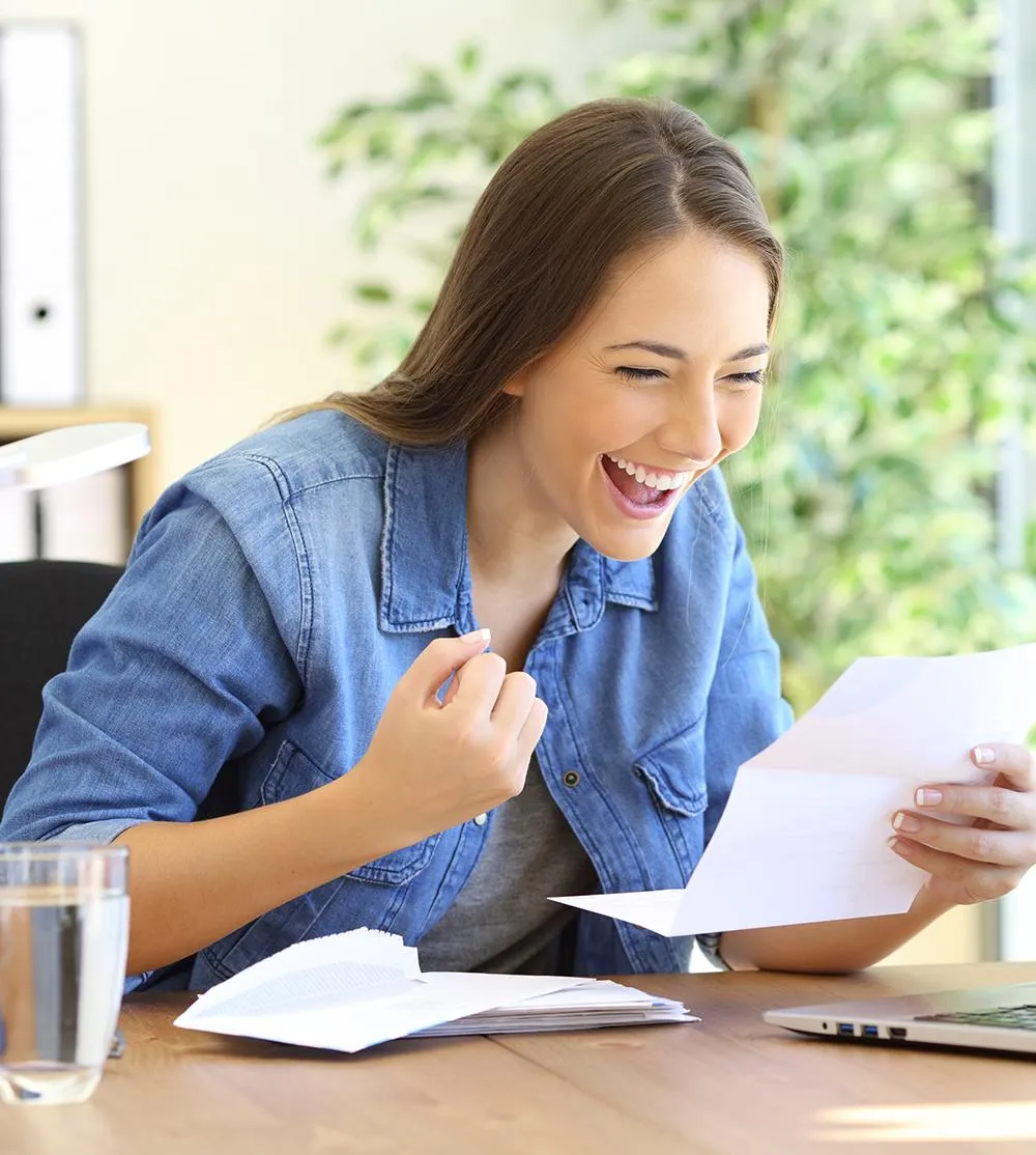 A woman joyfully laughs at her desk, engaged with her laptop, creating a lively and cheerful atmosphere