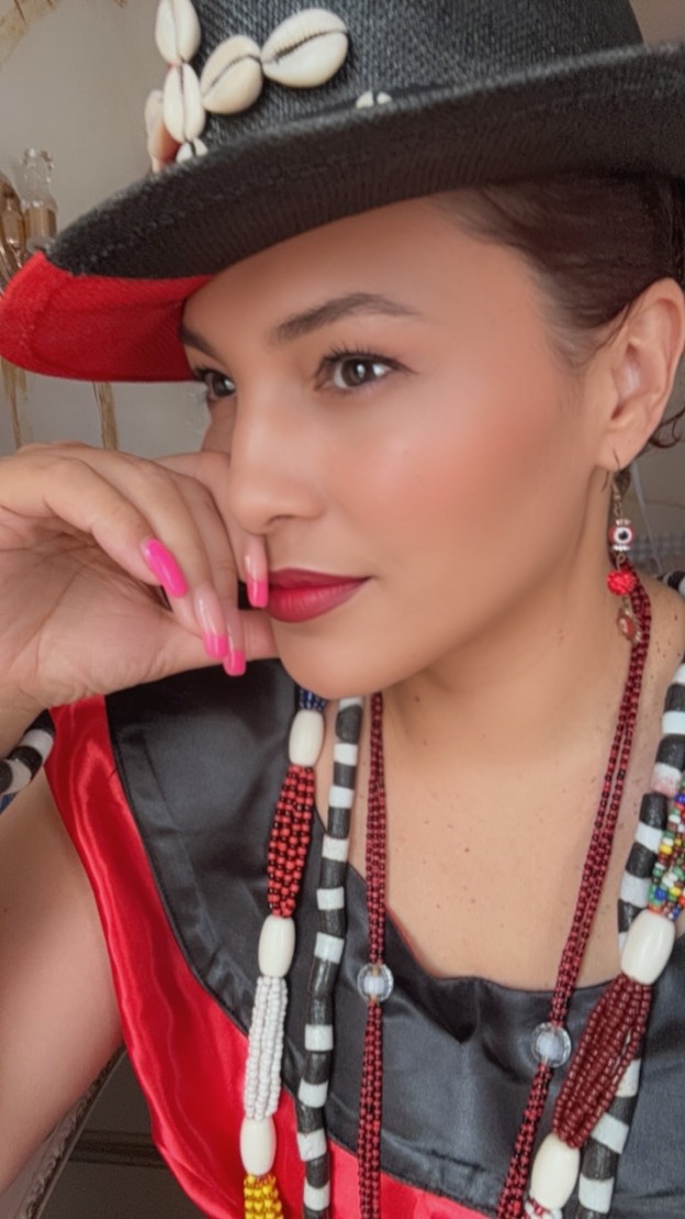 Close portrait of Omi in a black straw hat trimmed with cowrie shells, wearing layered red, white and black beaded strands.