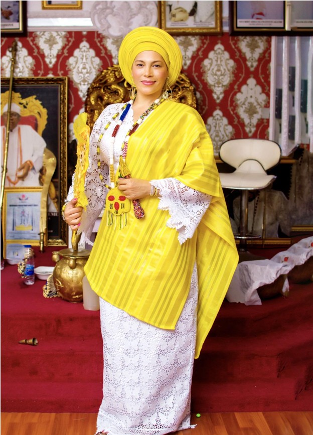 Omi in a yellow aso-oke gele and shawl over white lace, holding a gold staff in a formal reception room.