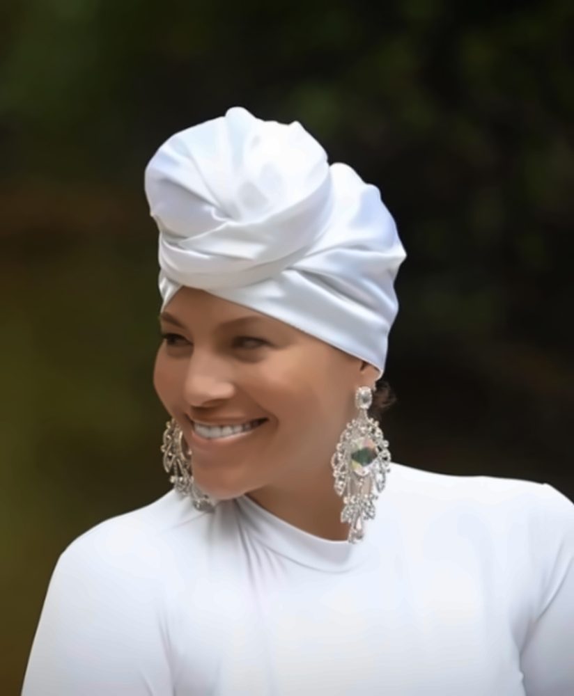 Close portrait of Omi smiling, in a white satin head wrap and white high-neck top with crystal chandelier earrings.