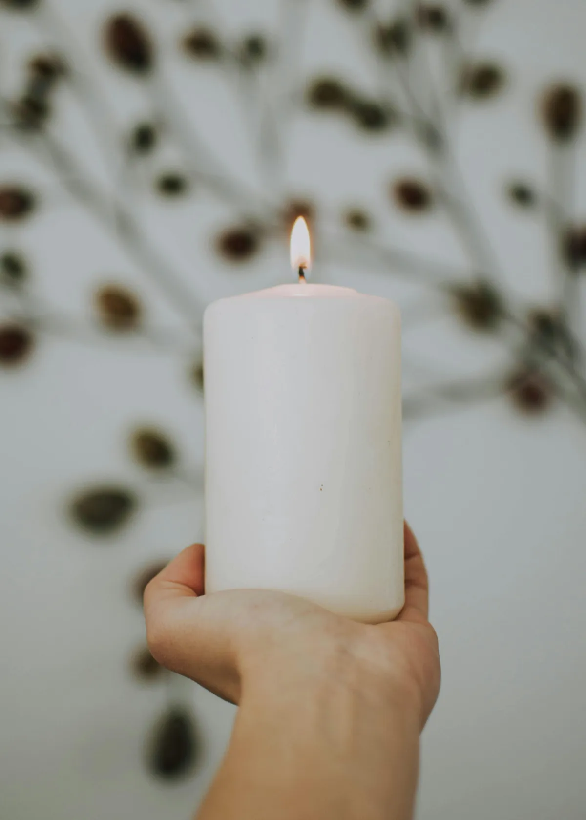 Close-up of hands holding a glowing white pillar candle against a softly blurred, neutral background.