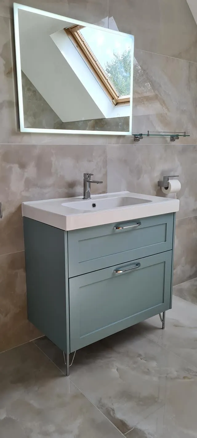 Modern bathroom in Meath featuring a light green vanity, illuminated mirror, and natural light from a skylight, complemented by neutral stone tiles.