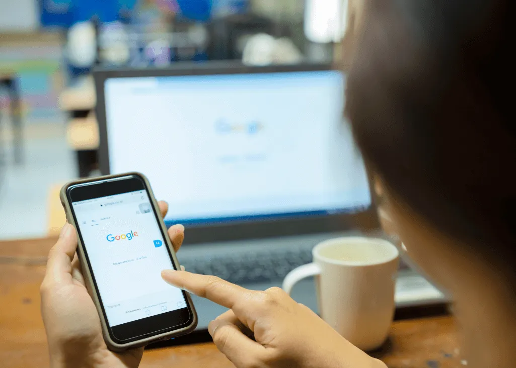 Person using smartphone to search on Google, with a laptop displaying Google search in background, emphasizing local business visibility for landscaping services.