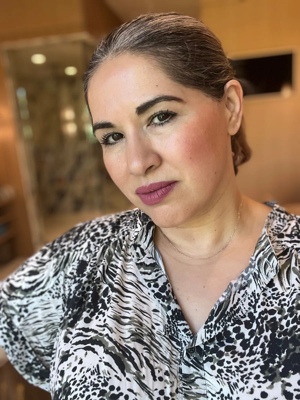 A woman with tied-back hair, wearing a patterned shirt, looks at the camera with a neutral expression in a softly lit indoor setting.