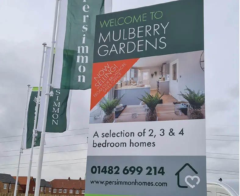 Large sign of Mulberry Gardens housing estate with green flags in the background and a view of the sky.