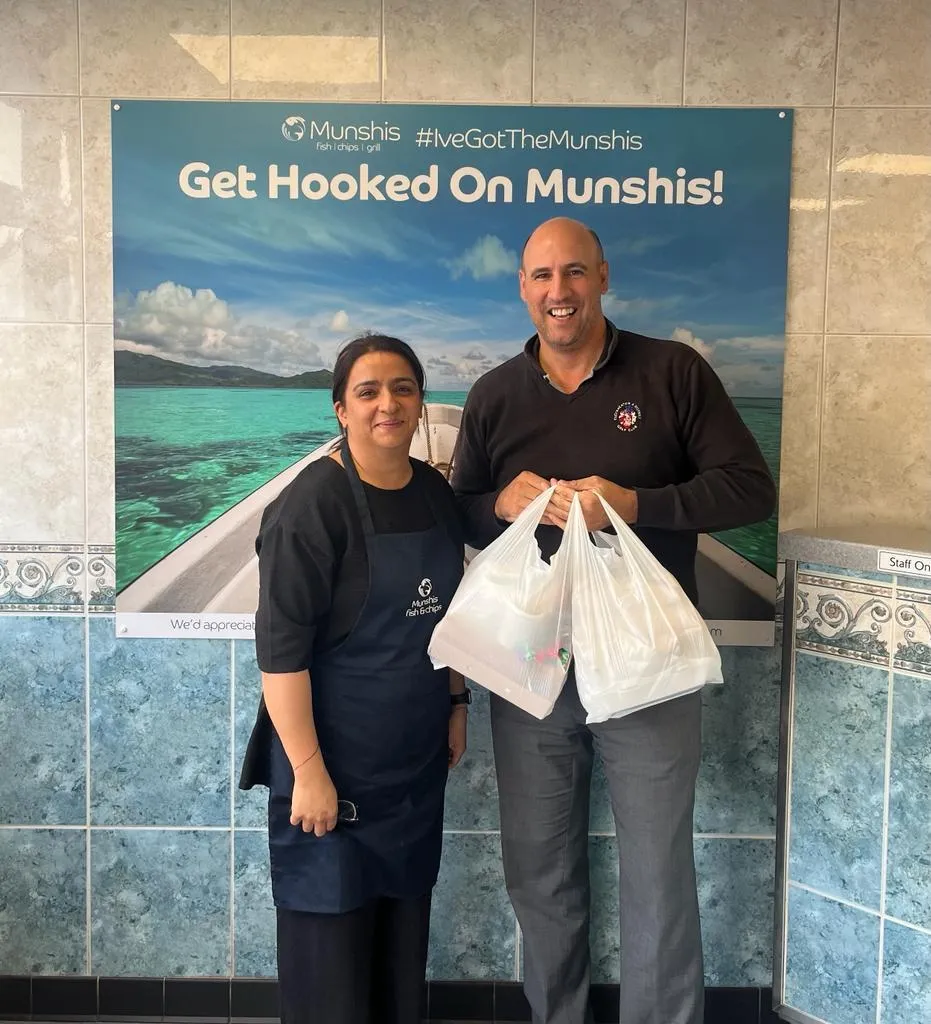 A male and female stood in front of a sign that says ‘Get Hooked on Munshis!’ inside a fish and chip shop. The male is holding two large white bags of food. 