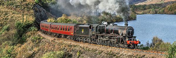 Red steamtrain on tracks with a view of a valley in the background with trees and foliage covering the surrounding areas. 