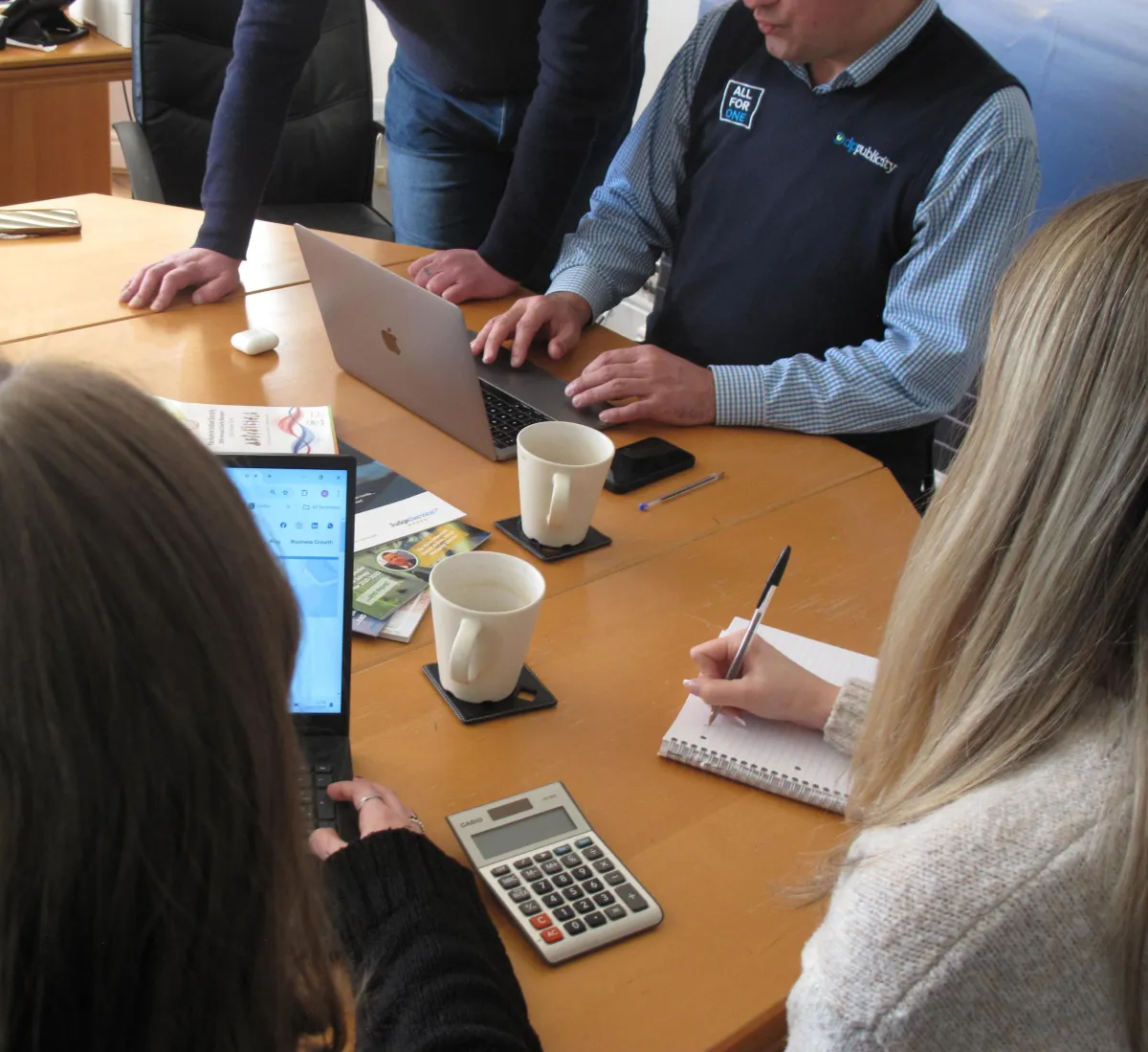 People in a business meeting, making notes and looking around a laptop screen with coffees on the desk.