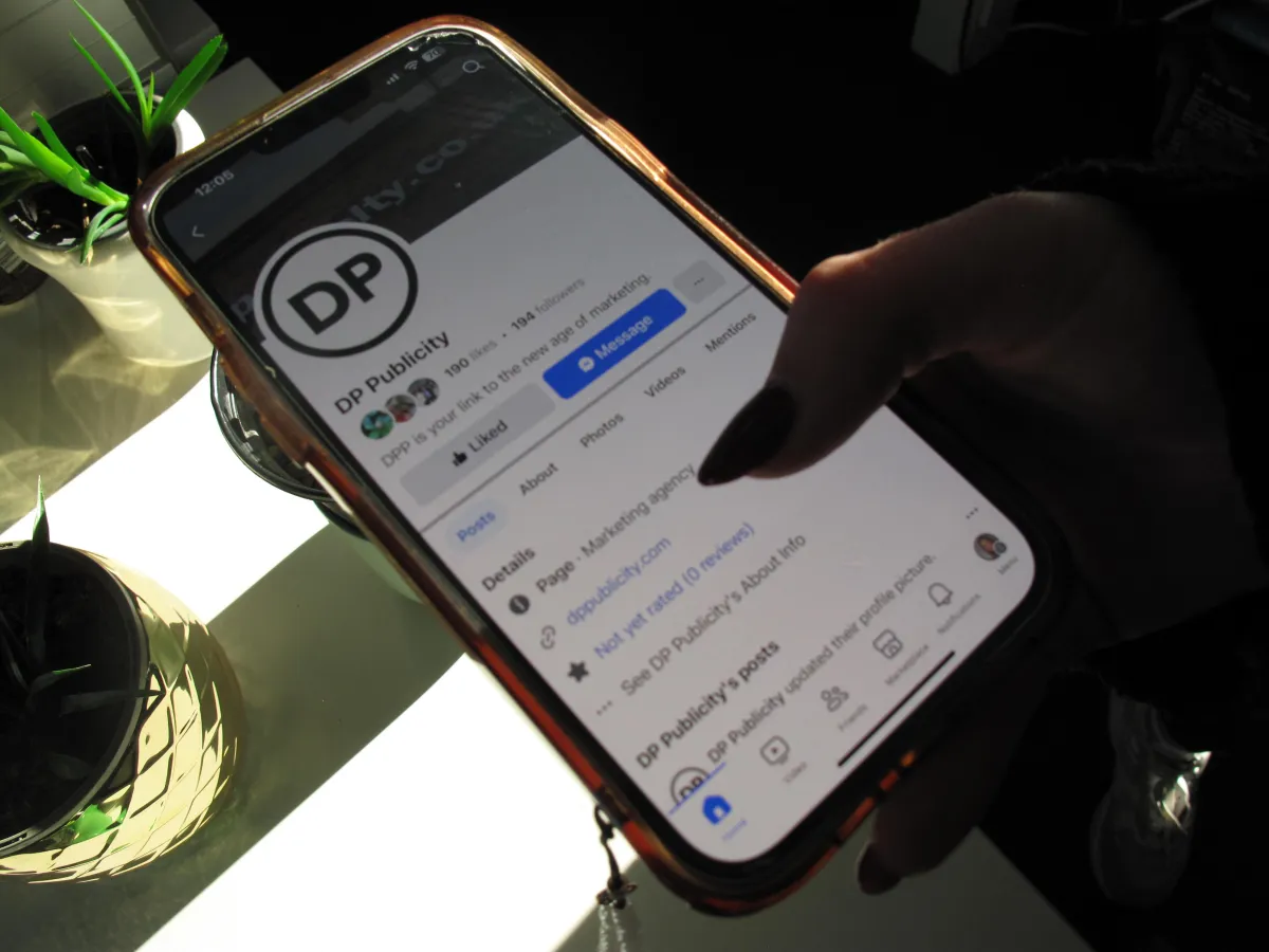 Hand holding a mobile phone with the DP Publicity Facebook page on screen with plants visible on a desk behind. 