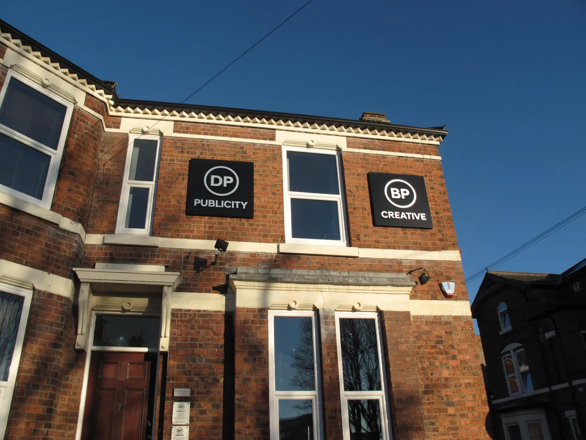 Image of the outside of the DP Publicity building with two large square signs on the upstairs section of the building. 