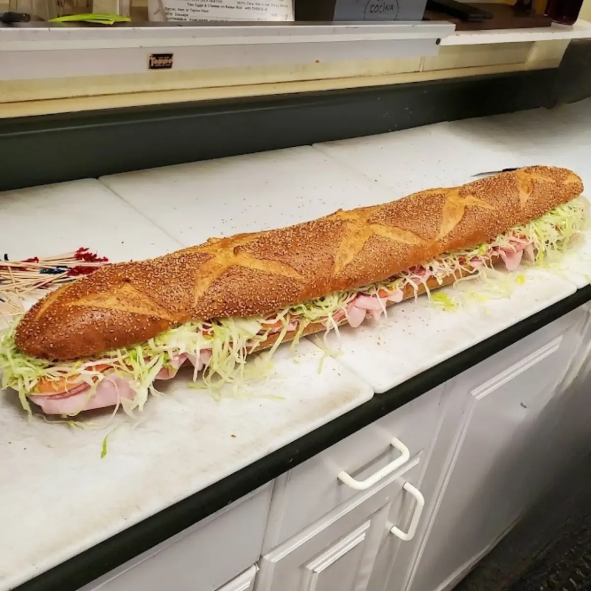 A massive, freshly made sub sandwich from La Cocina, filled with ham, cheese, shredded lettuce, and served on a crispy seeded baguette. Perfect for catering and large gatherings.