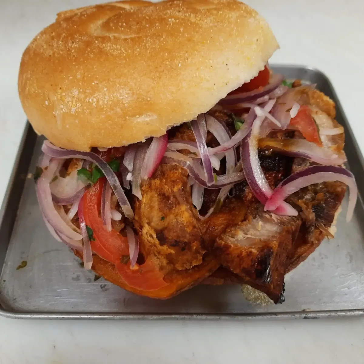 A delicious chicken sandwich with crispy pork, fresh tomatoes, and pickled red onions on a toasted bun, served on a metal tray at La Cocina.