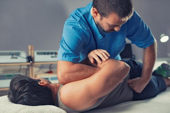 Man sitting down outdoors clutching his lower back in pain, wearing a blue shirt.