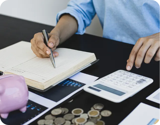 Hands writing in a notebook and using a calculator, with coins and a piggy bank, symbolizing saving time and money on marketing materials
