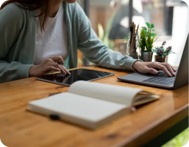 Close-up of a person's hands using a laptop and tablet on a wooden desk, with an open notebook and plants in the background.