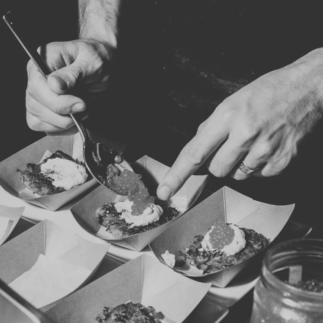 A chef's hands delicately garnishing small plates with a spoon, adding a dollop of cream and caviar to a gourmet dish, set against a dark background for H Brand