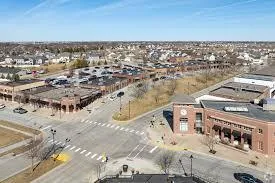 Wide aerial photo of a suburban commercial area with retail buildings, parking spaces, and surrounding residential neighborhoods under a clear blue sky.