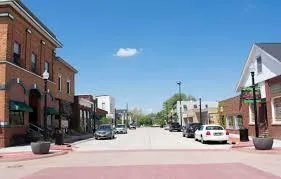 A small-town main street lined with brick buildings, shops, and parked cars under a bright blue sky.