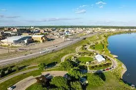 Aerial view of a waterfront park with walking paths, green spaces, and nearby commercial buildings under a bright blue sky.