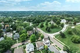 Aerial view of a suburban neighborhood with tree-lined streets, green lawns, and houses spread across rolling hills.