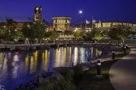 Nighttime view of a riverwalk area with illuminated buildings, reflections on the water, and pedestrians enjoying the evening scene.