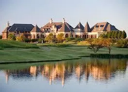 Elegant estate-style homes overlooking a lake and manicured golf course at sunset, with soft light reflecting on the water.