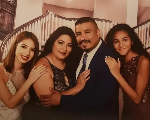 A smiling quinceañera family posing together indoors, with the parents embracing their daughters before the big celebration.