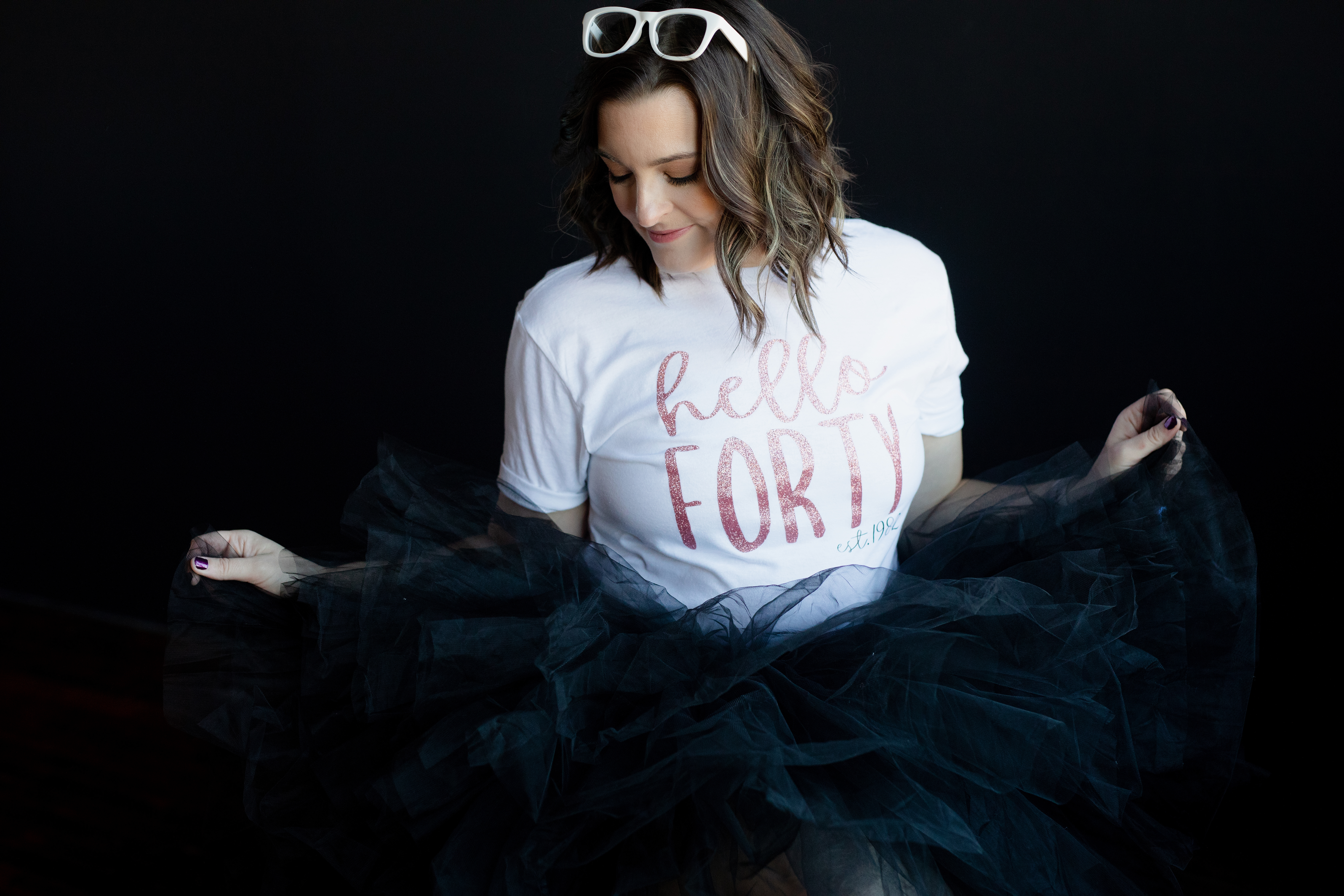 Creative birthday portrait of a woman in a “Hello Forty” shirt styled with a black tulle skirt in a dark studio setting.