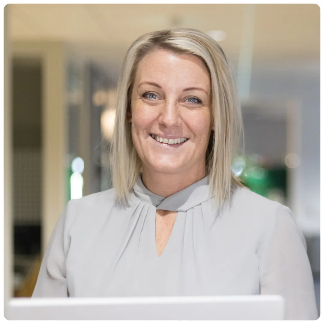 Profilbild av Sara Hellberg som ler mot kameran. Hon har en ljusgrå blus på sig och man skymtar en del av en datorskärm framför henne.