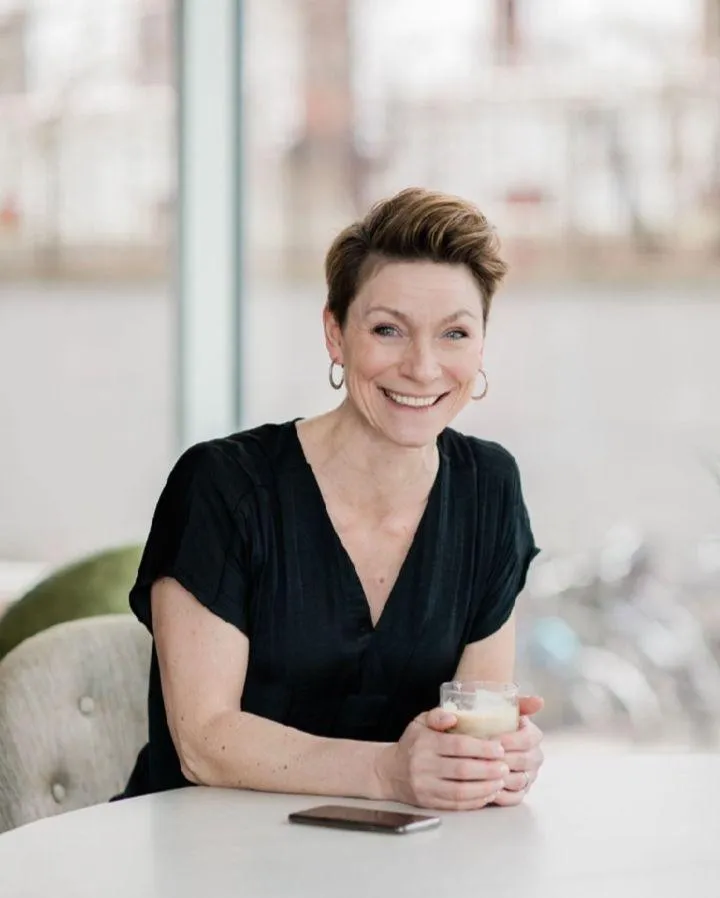Jill Nyqvist sitter vid ett bord och håller i ett glas. Hon ler mot kameran och på bordet ligger en telefon