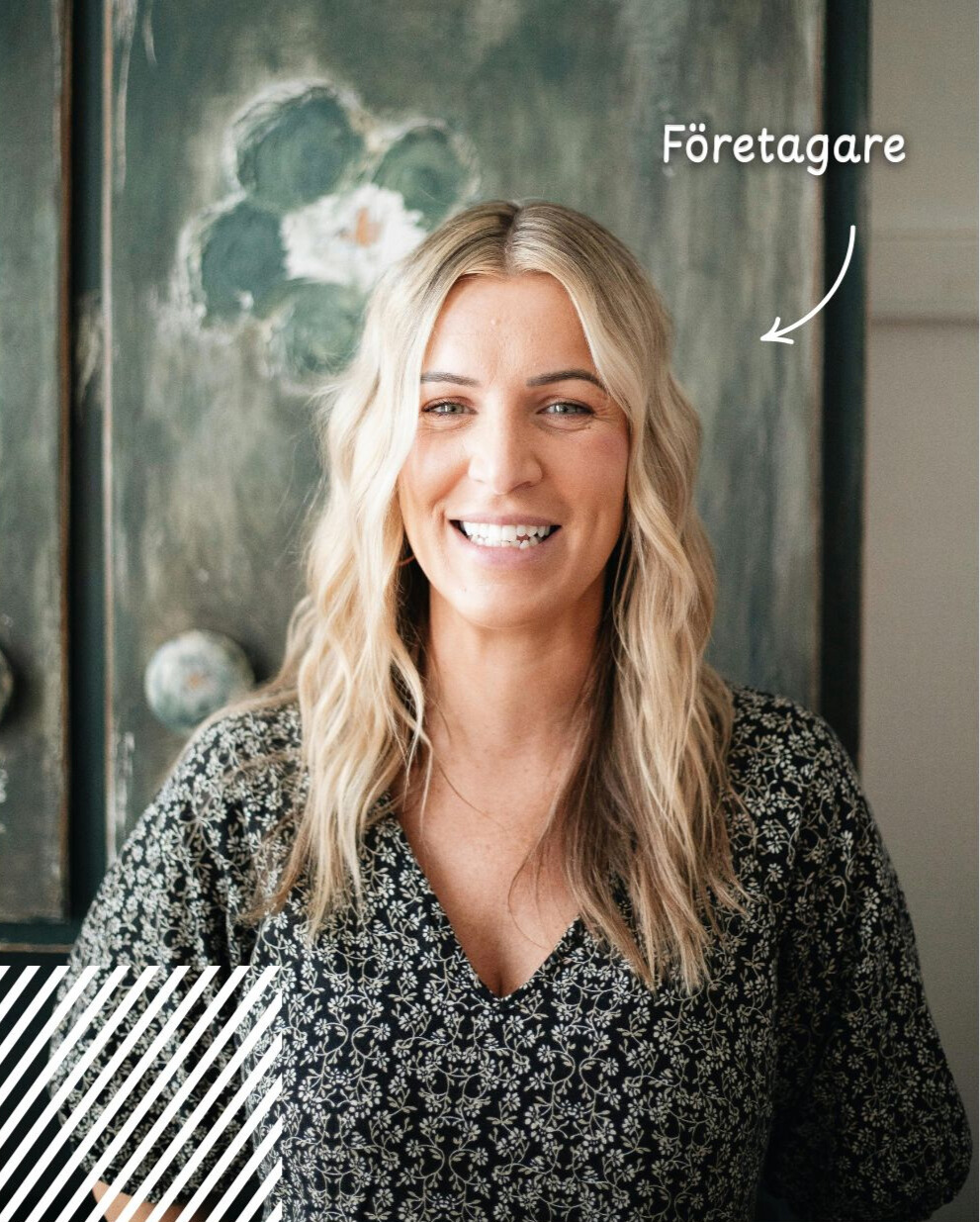 En blond småföretagare med en svart blus med vitt blommigt mönster ler mot kameran. I bakgrunden syns en tavla i akvarell i nyansen grönt