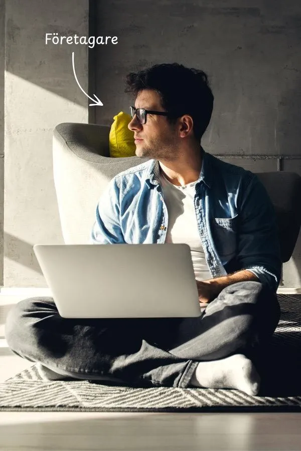 En manlig småföretagare sitter på golvet med en laptop i knät och tittar åt ena hållet. I bakgrunden syns en grå betongvägg, en grå soffa med en gul kudde och han sitter med benen i kors på en grå mönstrad matta.