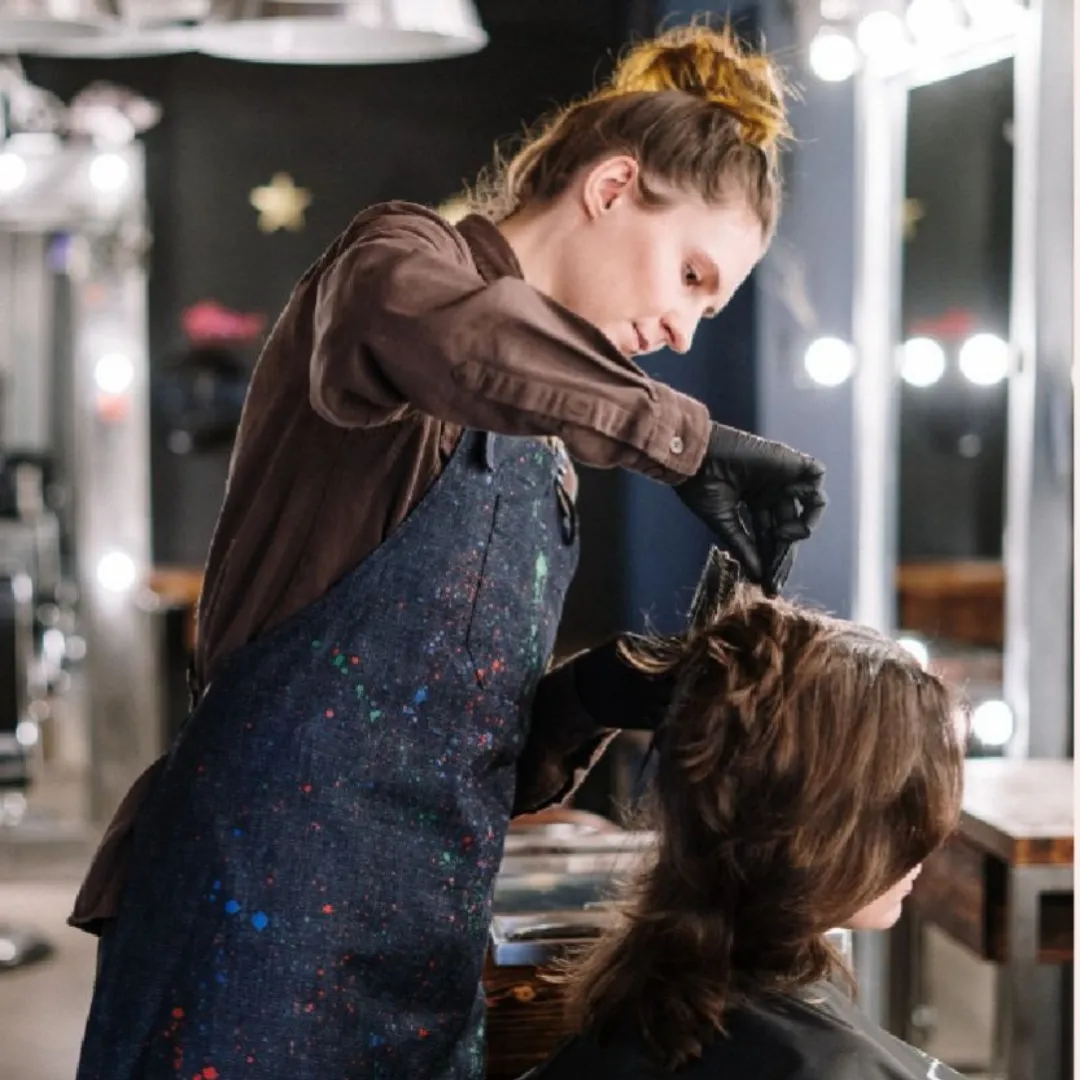 En frisör med håret uppsatt i en boll har på sig en brun skjorta, ett jeansfärgat förkläde med färgstänk och svarta engångshandskar klipper en kunds hår.