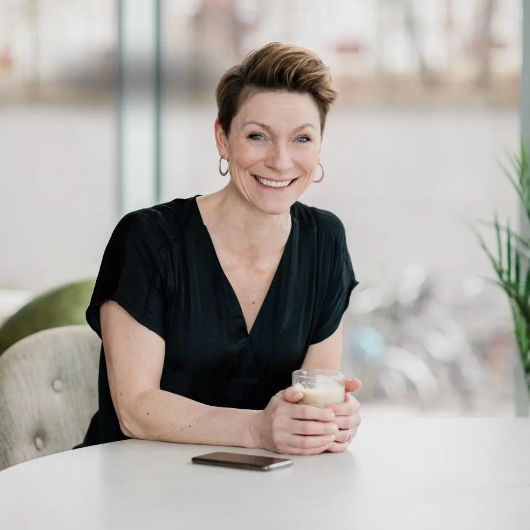 Jill Nyqvist på Affärskompetens - Företagarbyrån sitter vid ett bord och håller i ett glas. Hon ler mot kameran och på bordet ligger en telefon