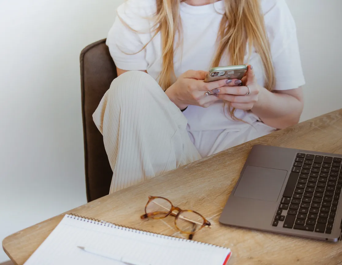 en blond kvinna som man inte ser ansiktet på sitter vid ett skrivbord och skriver på sin telefon. På skrivbordet står en bärbar dator, ett par glasögon samt ett block och en penna