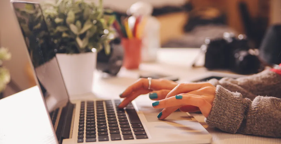 Närbild på en laptop från sidan där man ser ett par kvinnohänder med blått nagellack och en grå tjocktröja. I bakgrunden ser man en vit blomkruka med en grön växt, en systemkameran och en burk med pennor