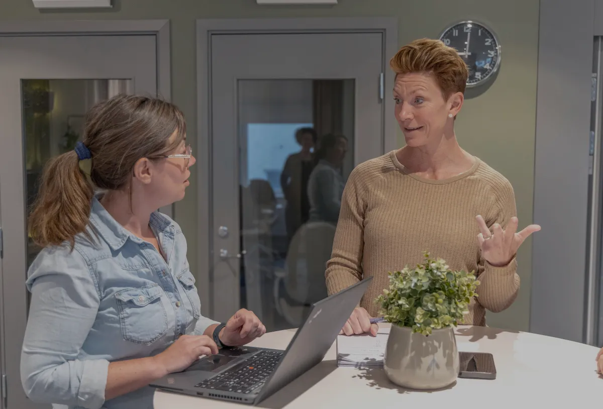 Företagsutvecklarna Hanna Rudolfsson och Jill NYqvist på Företagarbyrån står vid ett runt bort och diskuterar innehållet i Företagarklubben. Framför sig har de en öppen laptop och en blomkruka med en grön växt.