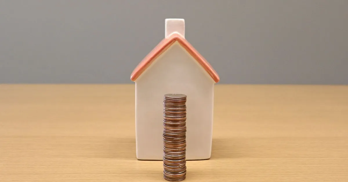 Small white ceramic house with a stack of coins in front of it on a wooden table