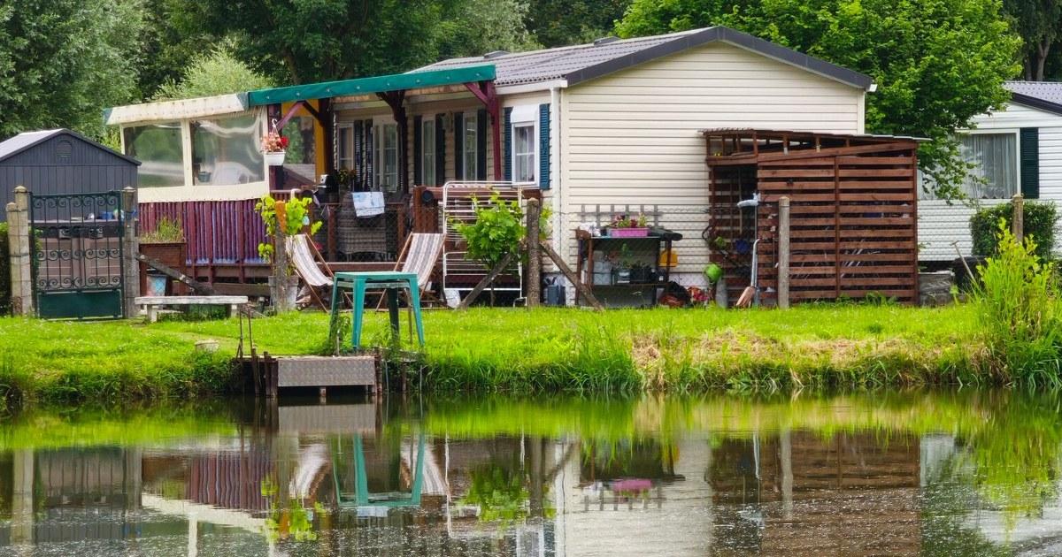 Cream manufactured home with a covered porch and wooden shed beside a calm pond with green trees behind it