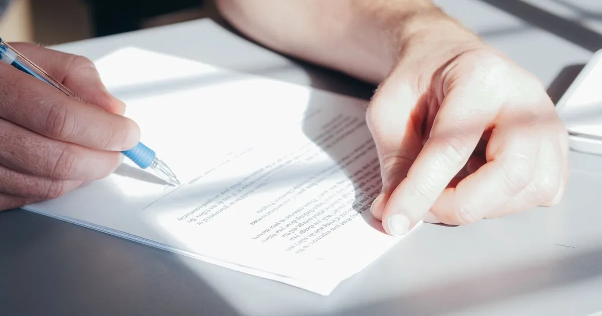 Two hands signing a printed document on a gray desk with a blue pen