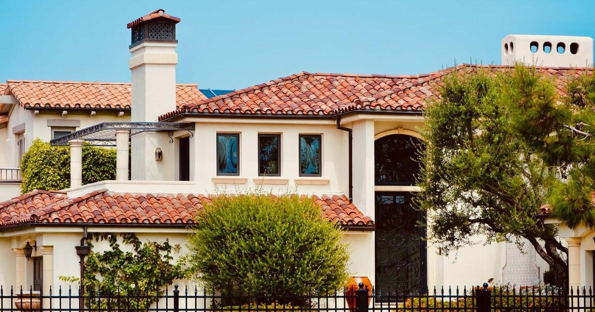 Two-story stucco home with a clay tile roof, green trees, and an iron fence